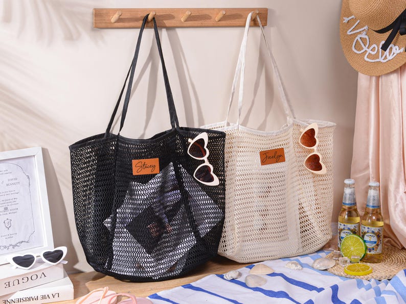 May include: Two mesh tote bags, one black and one off-white, with leather name tags and heart-shaped sunglasses. The black bag has "Stacey" on the tag, and the off-white bag has "Jocelyn". A straw hat and drinks are in the background.