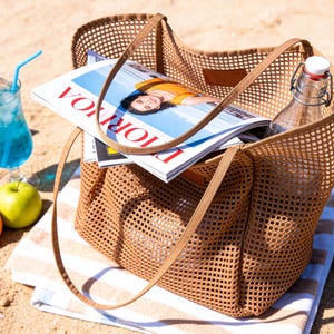May include: A brown mesh tote bag with long handles, filled with magazines, a glass bottle, and resting on a striped towel. A blue cocktail, watermelon, and fruit are nearby, suggesting a beach or outdoor setting.