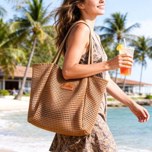 May include: A tan mesh tote bag with a leather patch that reads "Kristen". The bag has long handles and is carried by a person walking on a beach. The person is holding a drink with a straw and lemon slice.