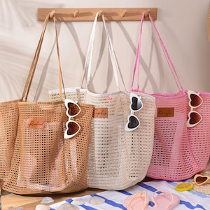 May include: Three mesh tote bags in tan, white, and pink, each with a long handle. Heart-shaped sunglasses are clipped to the bags. The bags have a rectangular leather patch with a name on them. The bags are on a beach-themed background.