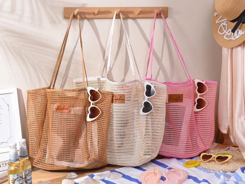 May include: Three mesh tote bags in tan, white, and pink, each with a long handle. Heart-shaped sunglasses are clipped to the bags. The bags have a rectangular leather patch with a name on them. The bags are on a beach-themed background.