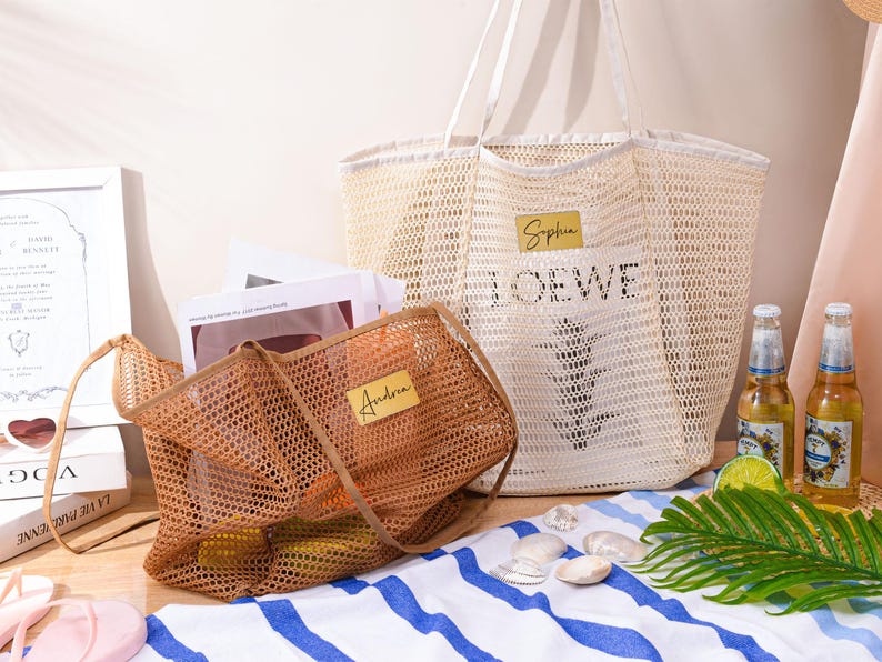 May include: Two mesh tote bags, one tan and one brown, with personalized name tags. The larger bag has "Sophia" and "LOEWE" printed on it. The bags are on a striped towel with seashells, drinks, and a palm leaf, suggesting a beach theme.