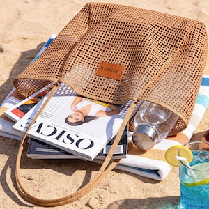 May include: A tan mesh tote bag with a leather tag that reads "Christine" sits on a beach towel with magazines and a water bottle inside. A glass of blue liquid with a lemon slice and a wooden tray with fruit are also visible.
