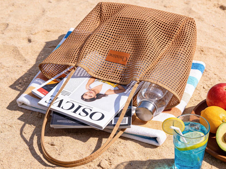 May include: A tan mesh tote bag with a leather tag that reads "Christine" sits on a beach towel with magazines and a water bottle inside. A glass of blue liquid with a lemon slice and a wooden tray with fruit are also visible.