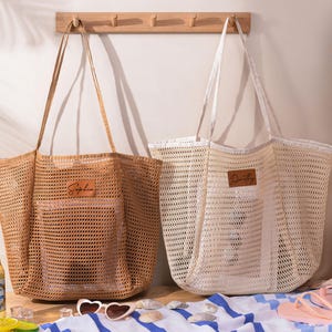 May include: Two large, open-weave tote bags. One is tan, the other is off-white. Each has a rectangular leather patch with a name on it. The bags have long handles and are designed for carrying items. The bags are on a striped towel.
