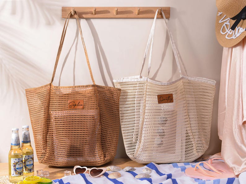 May include: Two large, open-weave tote bags. One is tan, the other is off-white. Each has a rectangular leather patch with a name on it. The bags have long handles and are designed for carrying items. The bags are on a striped towel.