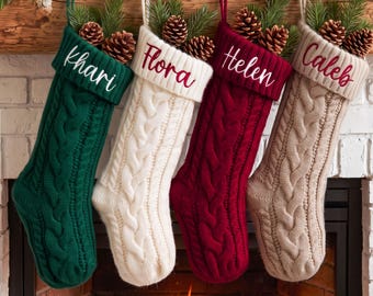 May include: Four cable-knit Christmas stockings in dark green, cream, deep red, and beige. Each stocking features a ribbed cuff embroidered with the names Khari, Flora, Helen, and Caleb respectively, and is topped with pine branches and pinecones.