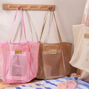 May include: Three mesh tote bags in pink, tan, and off-white, each with a name tag. The pink bag has the name "Sophia", the tan bag has "Amanda", and the off-white bag has "Hannah". The bags have long handles.