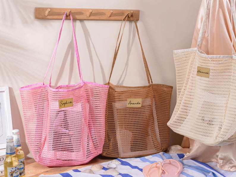 May include: Three mesh tote bags in pink, tan, and off-white, each with a name tag. The pink bag has the name "Sophia", the tan bag has "Amanda", and the off-white bag has "Hannah". The bags have long handles.