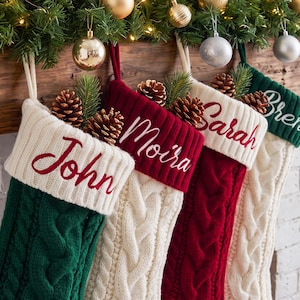 Op de afbeelding: Vier gebreide kerstsokken met kabelpatronen en witte boorden geborduurd met de namen John, Moira en Sarah. Elke sok is versierd met een dennenappel en een takje groen, hangend aan een houten schoorsteenmantel.