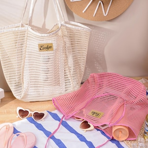 May include: A beige mesh tote bag with the name "Evelyn" on a gold label, and a pink mesh hat with the name "Bella" on a gold label. The items are on a striped towel with heart-shaped sunglasses and pink sandals.