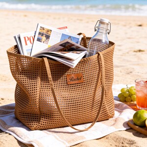 May include: A brown mesh tote bag filled with magazines and a glass water bottle, sitting on a beach towel. The bag has long handles and a leather tag that says "Rachel". Refreshments and fruit are arranged nearby.