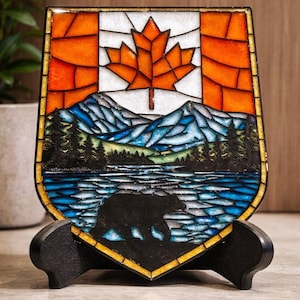 Op de afbeelding: Een gebrandschilderd schild met de Canadese vlag, een esdoornblad en een landschap. Het landschap omvat bergen, een meer, bomen en een berensilhouet. Het schild wordt op een zwarte standaard getoond.
