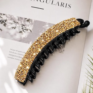 May include: A black banana hair clip decorated with glittering golden rhinestones. The clip is curved and has teeth for holding hair in place. The rhinestones are closely set, producing a sparkling appearance.