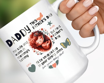 Op de afbeelding: Witte keramische mok met een afbeelding van een baby in de baarmoeder, omringd door bloemen en vlinders. De tekst luidt "Daddy This Father's Day I'm safe and warm in mommy's tummy but next Father's Day I'll be snuggled up in your strong arms Love, your Baby Girl!"