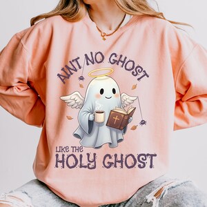 May include: A light pink sweatshirt with the phrase "Aint No Ghost Like The Holy Ghost" in purple. The graphic features a cartoon ghost with angel wings, a halo, a coffee cup, and a book.