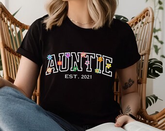 May include: Black t-shirt with white text that reads "AUNTIE EST. 2021" with colourful flower accents.