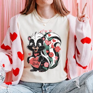 May include: A white t-shirt with a cartoon skunk holding a red heart that says "Love Stinks". The skunk is surrounded by pink and green flowers.