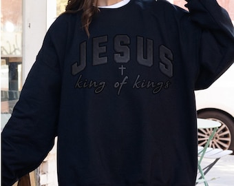 Könnte beinhalten: Schwarzes Sweatshirt mit dem Wort "JESUS" in erhabenen, dunklen Buchstaben, mit einem Kreuz und dem Schriftzug "king of kings" darunter. Das Sweatshirt hat einen Rundhalsausschnitt und lange Ärmel. Ein weißes Unterhemd ist sichtbar.