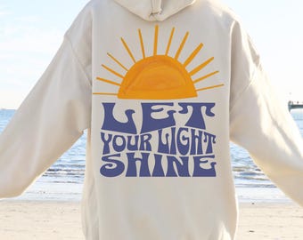 May include: A cream-colored hoodie with a yellow sun graphic and the text "LET YOUR LIGHT SHINE" in blue.