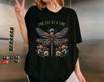 以下が含まれることがあります： 「ONE DAY AT A TIME」というテキストとトンボのグラフィックがプリントされた黒いTシャツ。トンボはカラフルな花でできています。