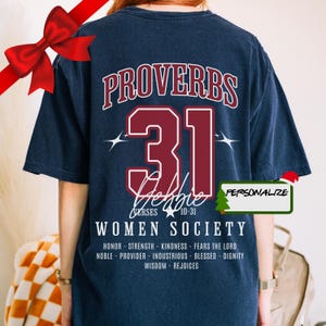 Puede incluir: Una camiseta azul marino con un gráfico rojo y blanco del texto "Proverbios 31" con una estrella y el texto "Sociedad de mujeres" debajo. El texto "Versículos 10-31" también está incluido en el gráfico. El texto "Personalizar" está en un cuadro verde en la esquina superior derecha de la imagen.
