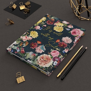 May include: A floral journal with a dark teal cover, adorned with a vibrant array of pink, yellow, and red flowers. The gold script reads "It is well with my Soul." Gold and black pencils and gold binder clips are also visible.