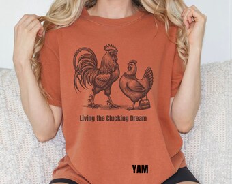 May include: A rust-colored t-shirt with a graphic of a rooster and a hen. The text "Living the Clacking Dream" is printed below the graphic. The word "YAM" is printed in the bottom right corner of the shirt.
