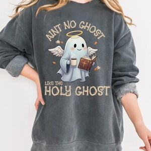May include: Dark gray sweatshirt with the text "Aint No Ghost Like The Holy Ghost" and a cartoon ghost design. The ghost is wearing a halo and holding a coffee cup and a book. The sweatshirt has long sleeves and a crew neck.