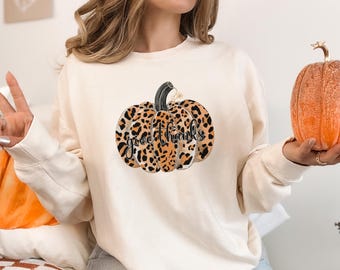May include: Cream-colored sweatshirt featuring a leopard print pumpkin graphic with the words "give thanks" in black script. The pumpkin has a black stem and is centered on the front of the long-sleeved top. A small orange pumpkin is held in the hand.