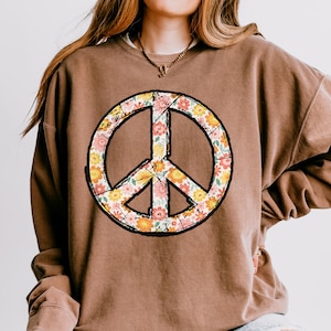 May include: A brown sweatshirt with a peace sign graphic. The peace sign is made up of a floral pattern in shades of pink, yellow, and orange.