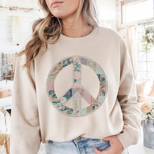 May include: Beige sweatshirt featuring a large peace sign. The peace sign is made of floral patchwork in shades of pink, blue, and green, with a fringed border. The sweatshirt has long sleeves and a crew neck.