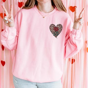 Puede incluir: Una sudadera rosa con un gráfico en forma de corazón que presenta un estampado de leopardo y una rosa roja. El corazón está centrado en el pecho de la sudadera.