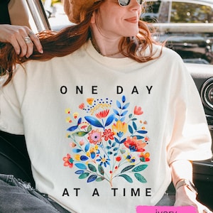 May include: A white long-sleeved t-shirt with a colorful floral design and the text "ONE DAY AT A TIME" in black. The shirt is ivory.