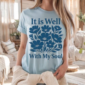 May include: Light blue t-shirt with a floral design and the text "It is Well With My Soul" in navy blue.