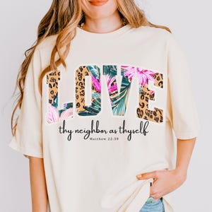 Op de afbeelding: Crèmekleurig T-shirt met het woord "LOVE" in grote, vette letters. De letters zijn versierd met een luipaardprint en tropisch bloemenontwerp. Onder het woord "LOVE" staat de zin "thy neighbor as thyself" en de verwijzing "Matthew 22:39".