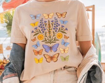 May include: A beige t-shirt with a floral design featuring butterflies in shades of blue, yellow, and orange. The butterflies are arranged around a cluster of purple and yellow flowers.
