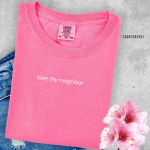 Op de afbeelding: Roze T-shirt met de tekst "love thy neighbor" in witte letters. Het shirt is gemaakt van een zacht, comfortabel materiaal.