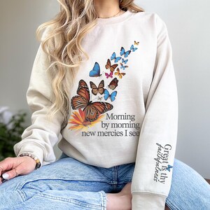May include: A beige sweatshirt with a colorful butterfly design and the text "Morning by morning new mercies I see" on the front. The right sleeve has the text "Great is thy faithfulness" with a small butterfly.