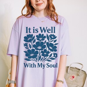 May include: A lavender t-shirt with a blue floral design and the text "It is Well With My Soul".
