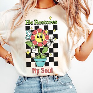 May include: A white t-shirt with a graphic design of a cartoon flower in a pot. The flower has a smiling face and is holding a musical note. The text "He Restores My Soul" is printed below the flower.
