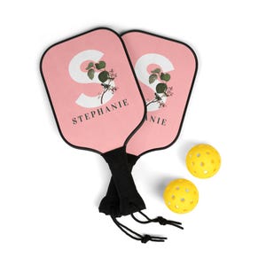 Könnte beinhalten: Zwei rosa Paddleball-Schläger mit schwarzen Griffen und einem floralen Design. Die Schläger haben den Buchstaben "S" und den Namen "STEPHANIE" aufgedruckt. Zwei gelbe Paddleball-Bälle sind enthalten.