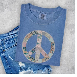 Puede incluir: Una camiseta azul con un símbolo de paz hecho de tela de patchwork. El símbolo de paz está en tonos de rosa, verde y marrón. La camiseta tiene una etiqueta que dice "Comfort Colors".