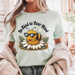 May include: A light blue t-shirt featuring a cartoon bee wearing sunglasses and sitting on a daisy. The text "Be Kind to Your Mind" is written in a retro font around the bee.