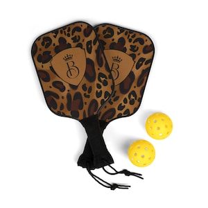 Könnte beinhalten: Zwei braune und schwarze Pickleball-Schläger mit Leopardenmuster, schwarzen Griffen und einem schwarzen Kordelzugbeutel. Zwei gelbe Pickleball-Bälle sind enthalten.