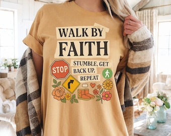 Puede incluir: Camiseta de color amarillo mostaza con el texto "WALK BY FAITH" en letras negras. La camiseta presenta un gráfico con una señal de stop, una señal de tráfico, botas, flores y las palabras "STUMBLE, GET BACK UP, REPEAT".