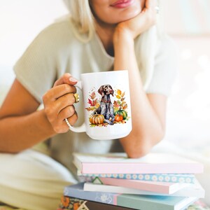 May include: White ceramic mug with a brown and white German Shorthaired Pointer dog sitting with pumpkins and fall leaves. The dog is surrounded by orange and yellow pumpkins and red and orange leaves.