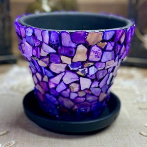 May include: Purple mosaic flower pot with a black saucer. The pot is made of small, irregular pieces of purple and white glass.