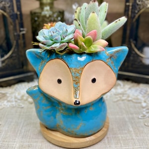 May include: A turquoise ceramic fox planter with a wooden base, filled with a variety of succulents. The fox planter has tan-colored ears and face, with black eyes and a small nose. The planter is a decorative item.