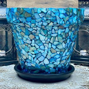 May include: A decorative flower pot with a mosaic design in shades of blue, teal, and white. The pot sits on a black saucer. Two black metal lanterns with lit candles flank the pot, creating a serene ambiance.